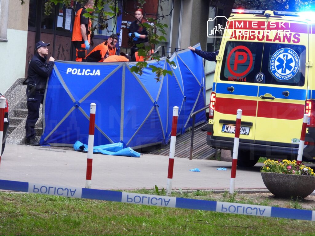 Policjant zabezpiecza miejsce zdarzenia, podczas gdy ratownicy medyczni pracują obok niebieskiego namiotu.