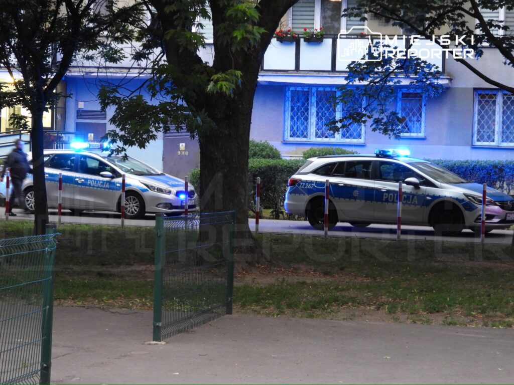 Dwa radiowozy policyjne z włączonymi sygnalizatorami świetlnymi stoją na osiedlowej ulicy.