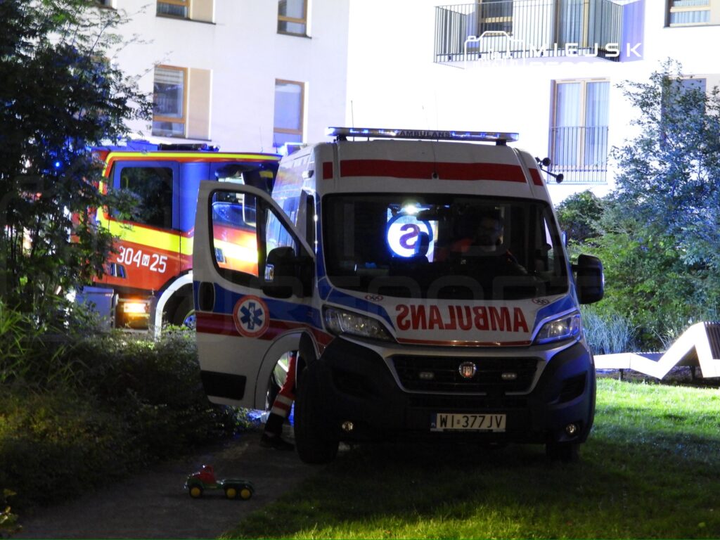 Ambulans z włączonymi sygnałami świetlnymi zaparkowany w ciemnym otoczeniu, obok wozu strażackiego.