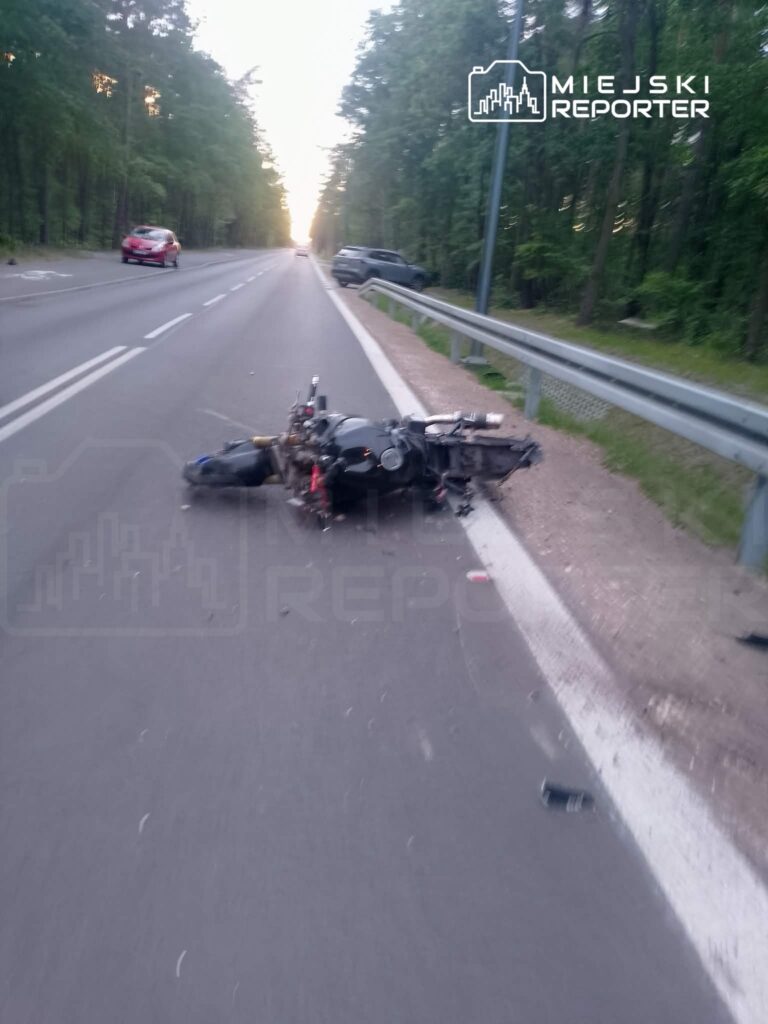 Motocykl leży na jezdni przy drodze otoczonej lasem, w tle widać zaparkowane samochody.