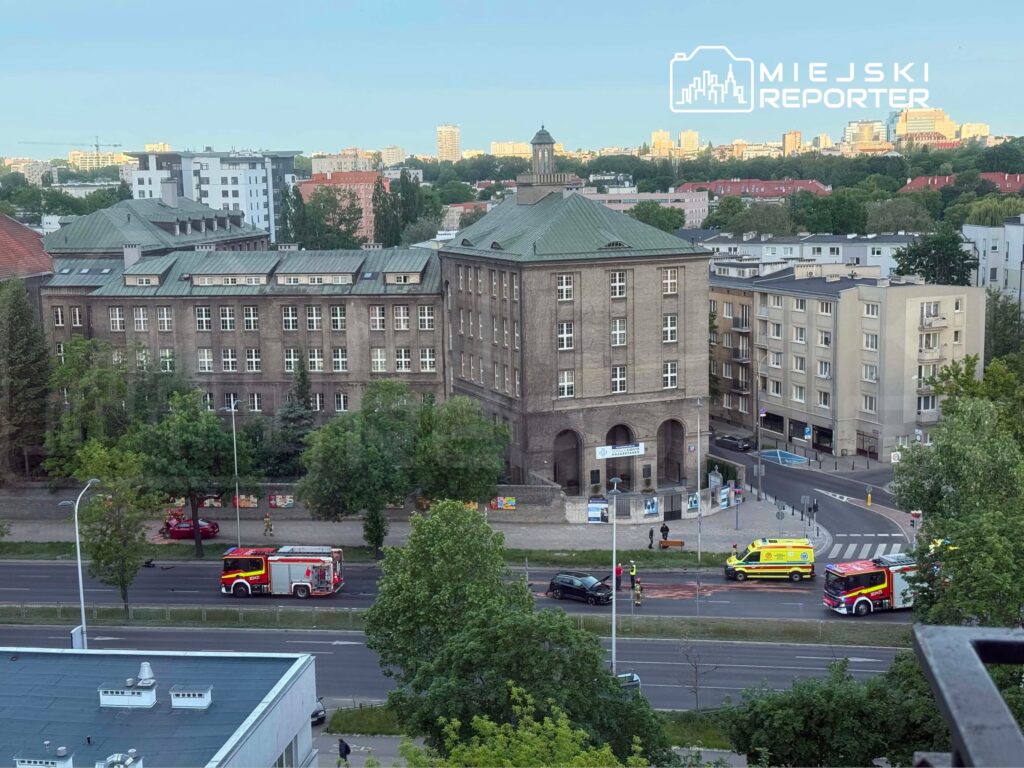 W tle nowoczesne budynki, na pierwszym planie widać straż pożarną i karetkę przy miejscu zdarzenia.