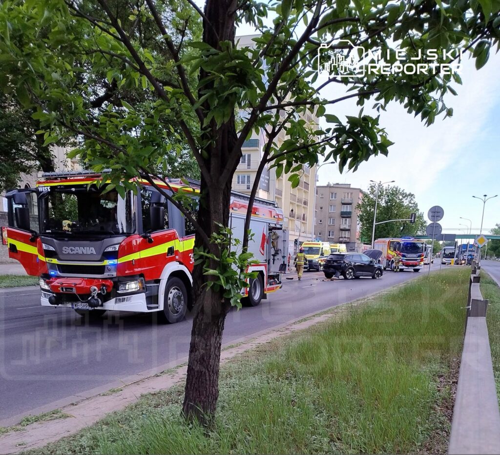 Strażacy pracują przy wypadku drogowym, obok uszkodzonego samochodu, na ruchliwej ulicy otoczonej budynkami mieszkalnymi.