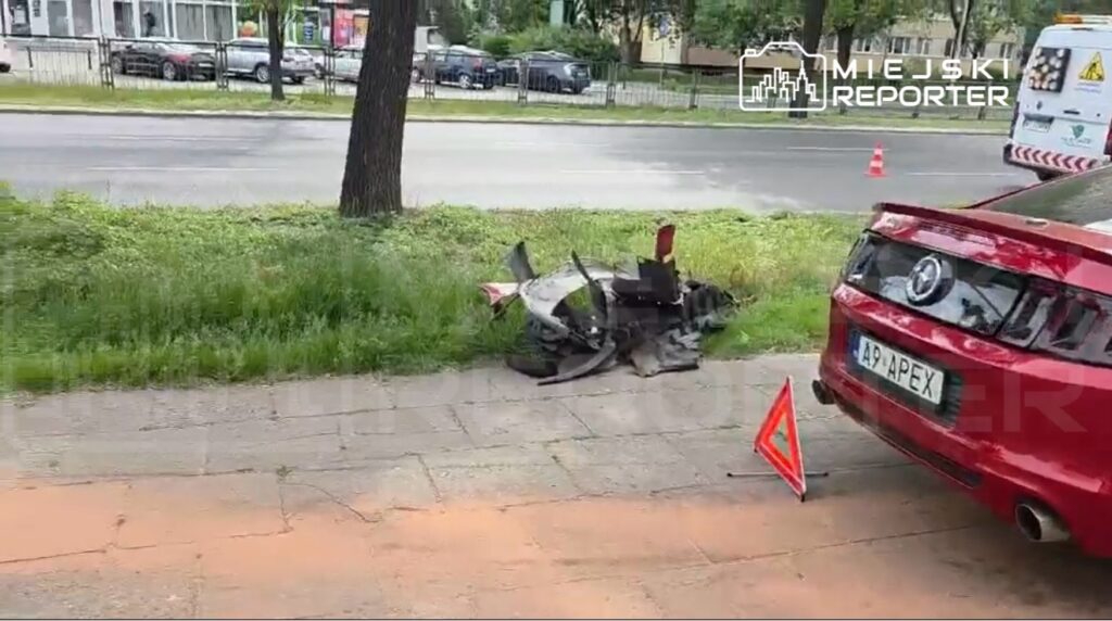Zniszczony fragment pojazdu leży na trawie obok ulicy, a w tle widać zaparkowane samochody oraz czerwoną osobówkę z trójkątnym znakiem ostrzegawczym.