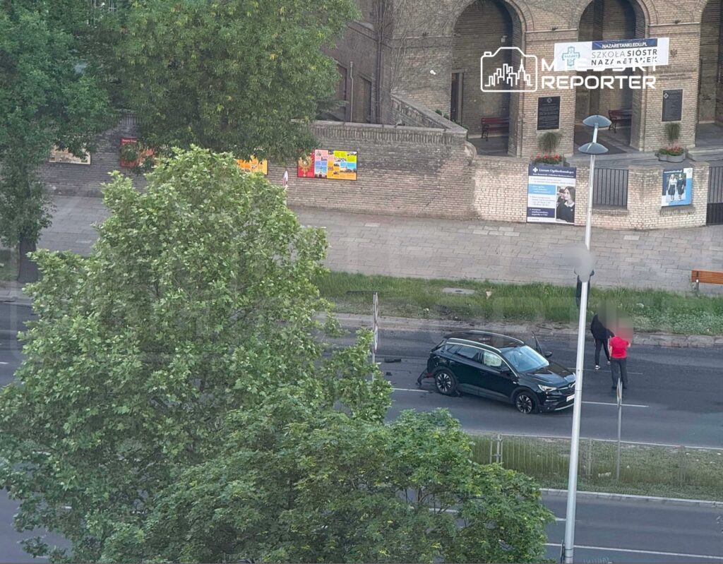 Czarny samochód zatrzymany na jezdni, obok dwóch osób rozmawiających w pobliżu budynku z plakatami.