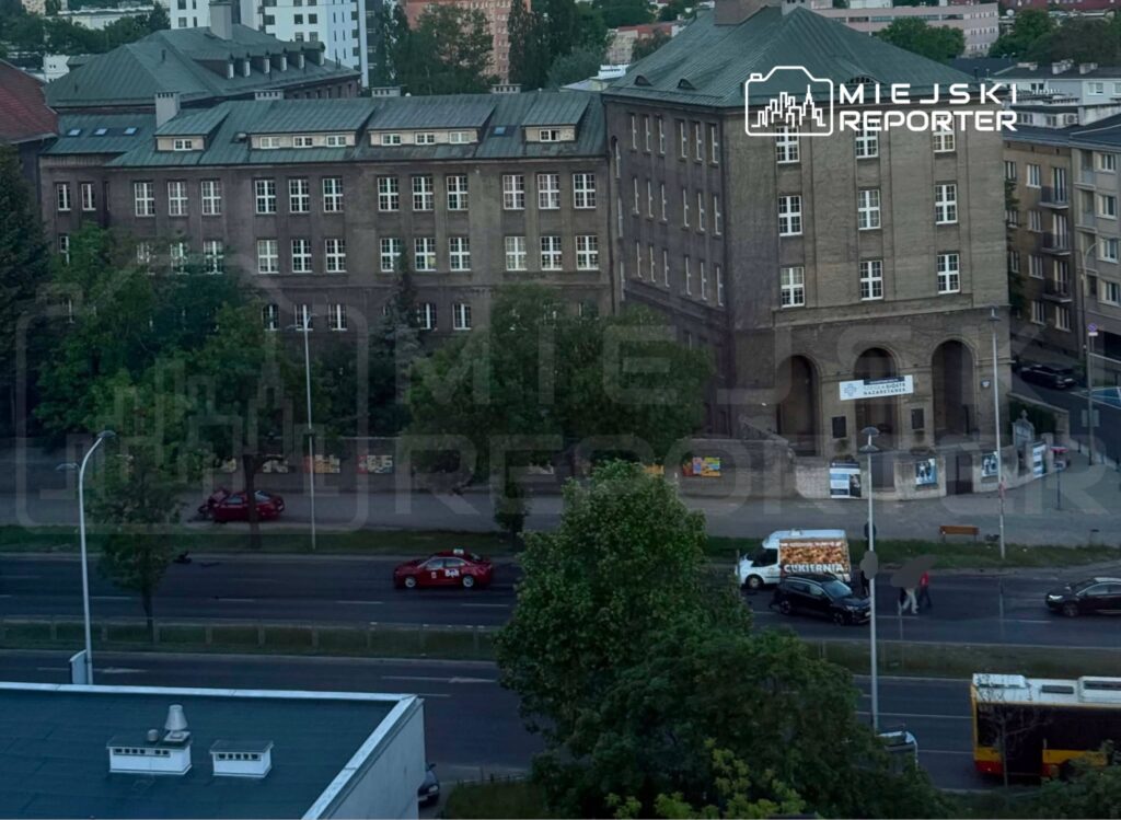 Czerwony samochód osobowy zatrzymany przy chodniku obok budynku z cegły, na którym widoczne są plakaty.