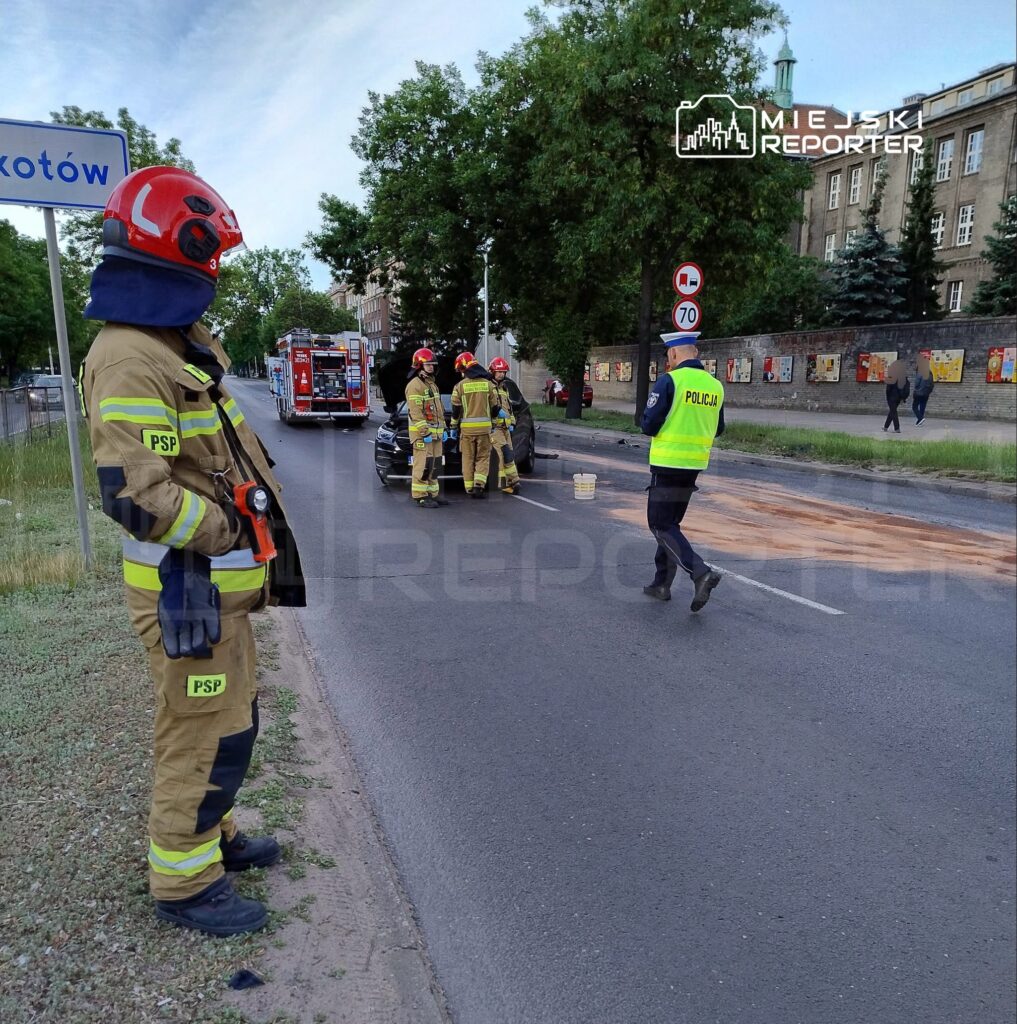 Strażacy w mundurach pracują przy uszkodzonym pojeździe na ulicy, obok nich policjant w kamizelce odblaskowej.