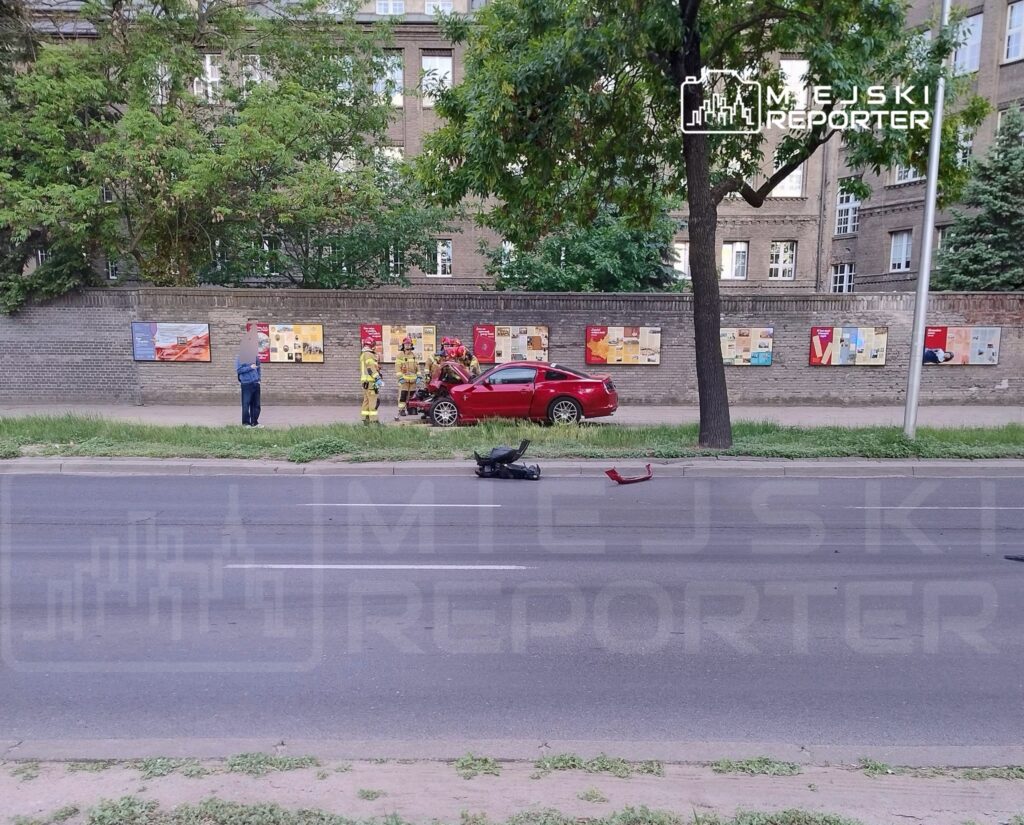 Grupa strażaków bada uszkodzony czerwony samochód na poboczu, obok leży uszkodzony skuter, w tle ściana z plakatami.
