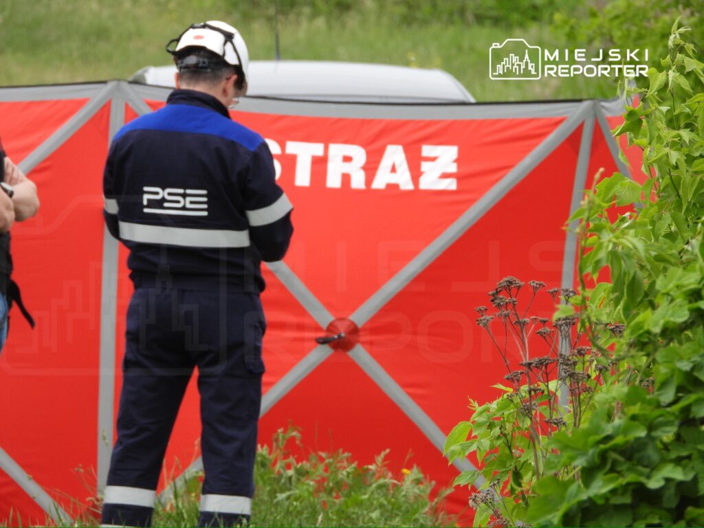 Mężczyzna w mundurze strażackim z logo PSE stoi przy czerwonym namiocie z napisem "STRAŻ", otoczony roślinnością.