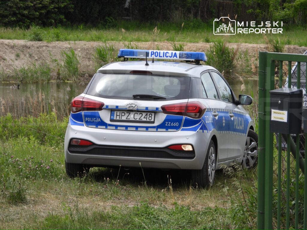 Policyjny samochód zaparkowany na trawie obok wody, w otoczeniu roślinności.