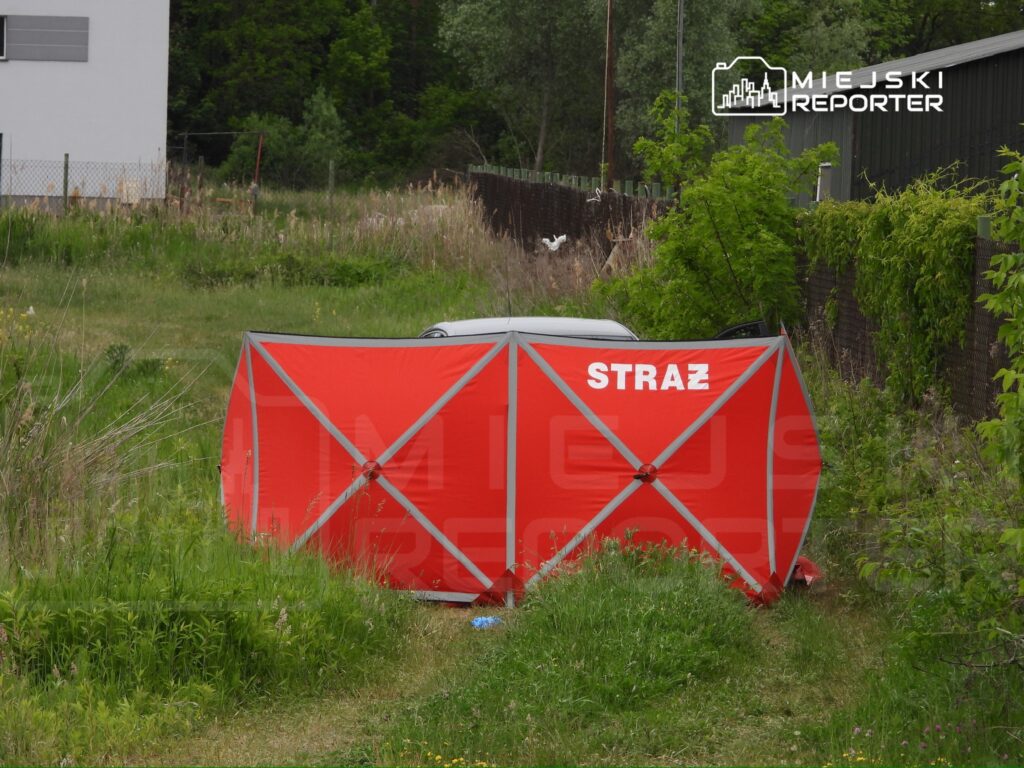 Czerwony namiot z napisem "STRAŻ" ustawiony na trawie obok zaparkowanego samochodu w zaroślach.