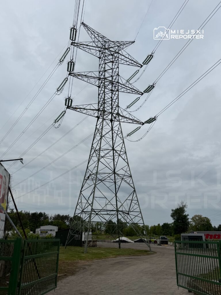 Wysoka wieża energetyczna z metalowej konstrukcji, otoczona zielenią, w pobliżu drogi i budynków gospodarczych.