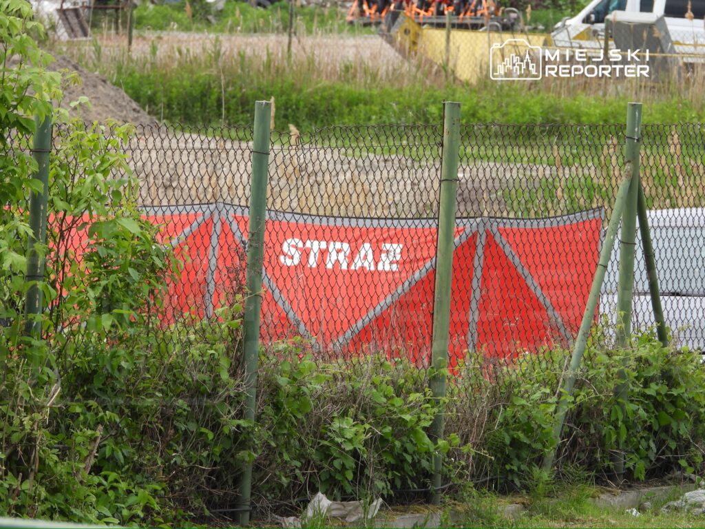Czerwony namiot z napisem "STRAŻ" ustawiony za ogrodzeniem, w otoczeniu zieleni i placu budowy.