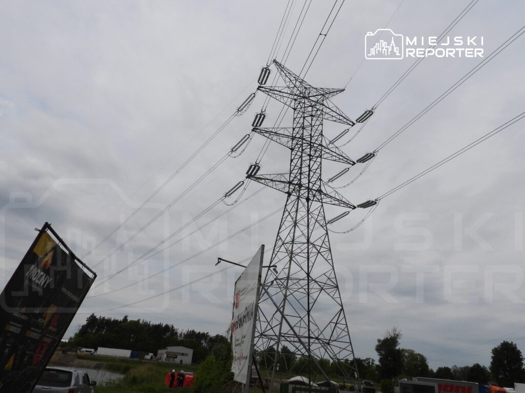 Wysoka wieża energetyczna z przewodami elektrycznymi, w tle chmurne niebo i zieleń otaczającego terenu.