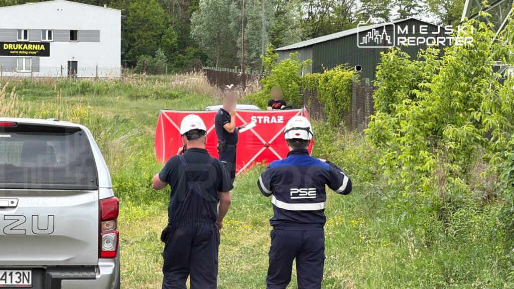 Grupa pracowników w kaskach, w tym mężczyzna z notatnikiem, omawia sytuację przy czerwonym namiocie z napisem "STRAŻ" w terenie zielonym.