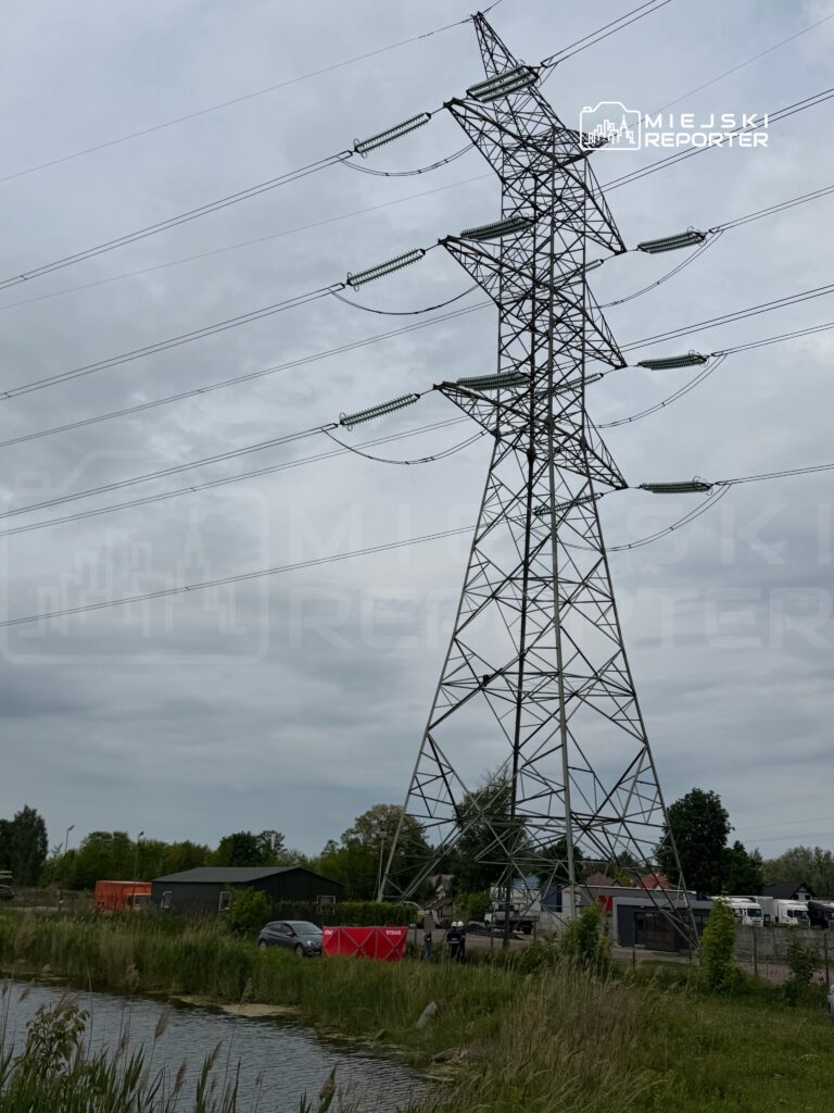 Wysoka wieża energetyczna z przewodami, w tle tereny zielone i budynki, przy brzegu wody.
