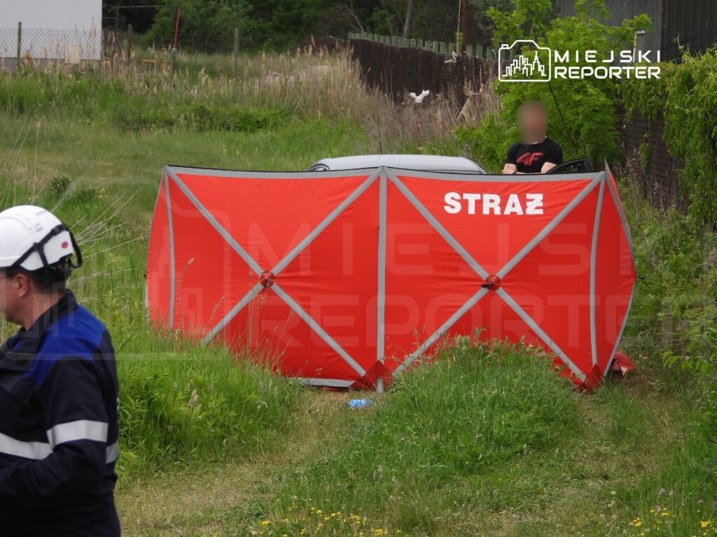 Strażacy w mundurach pracują w terenie przy czerwonym namiocie, obok samochodu w trawie.