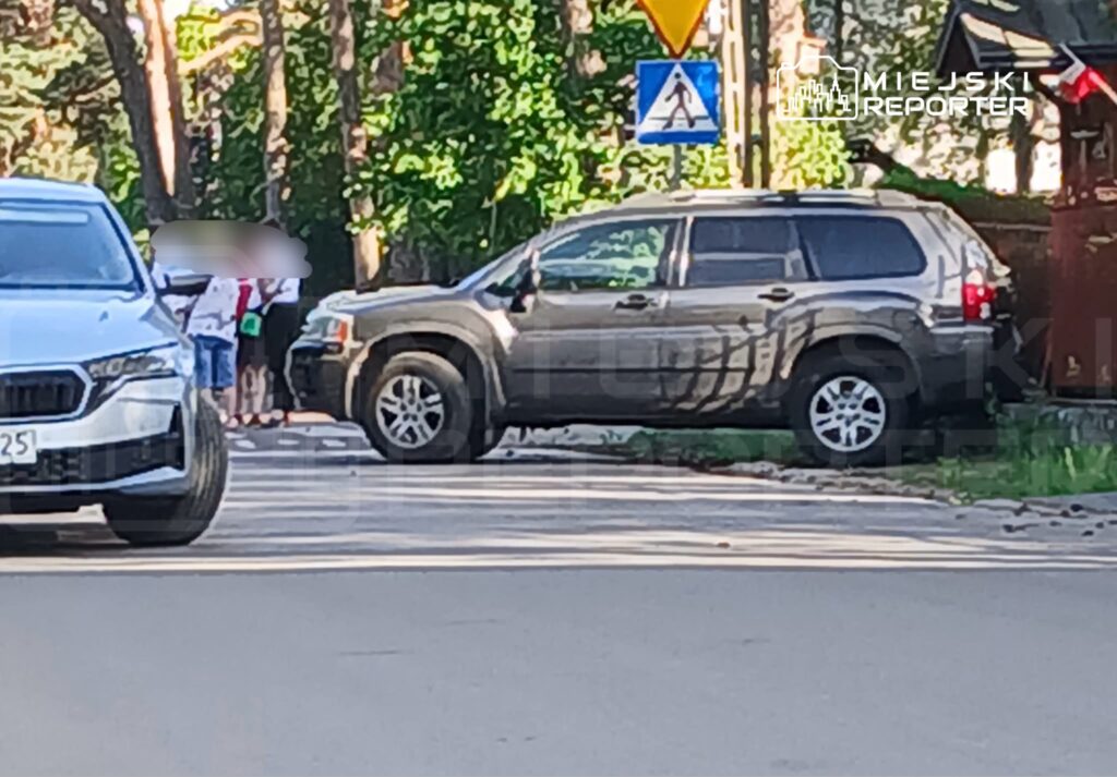 Czarny SUV stoi na poboczu drogi, obok grupy osób w tle, w otoczeniu drzew i znaków drogowych.