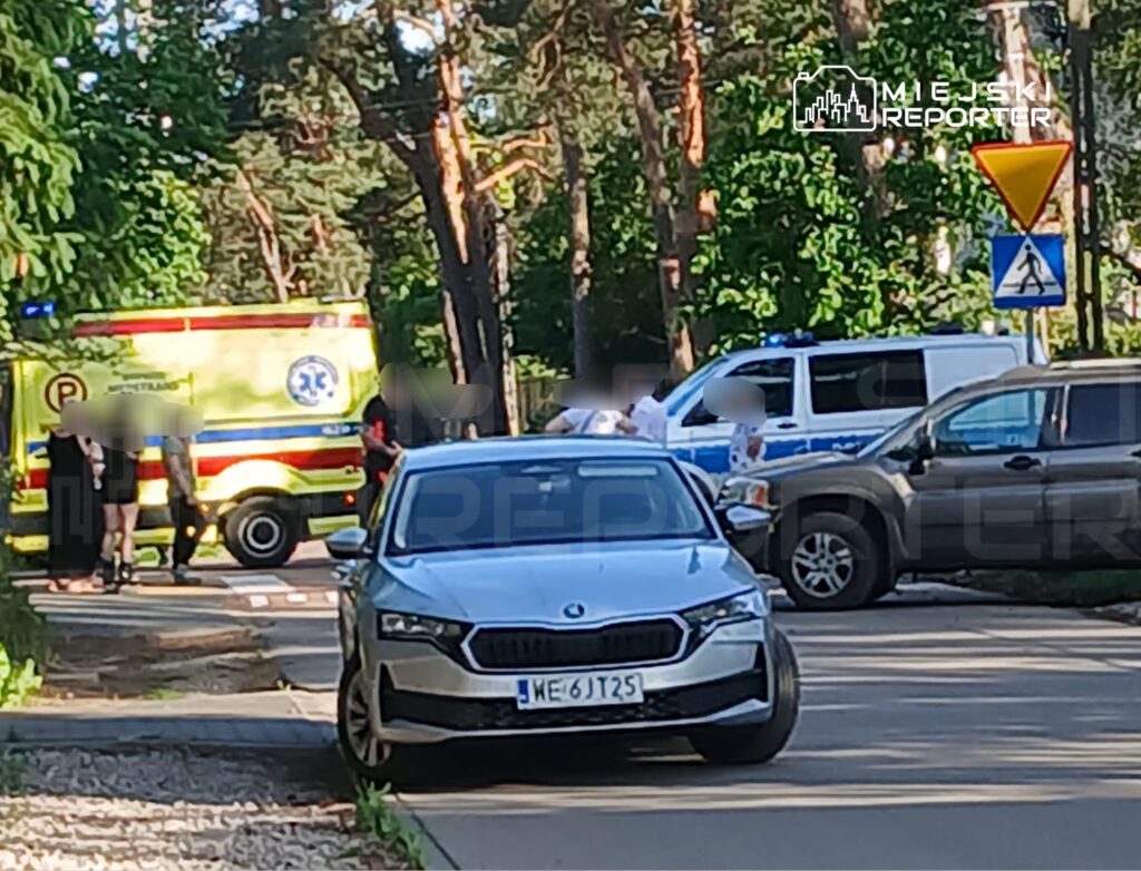 Karetka pogotowia oraz radiowóz policyjny zatrzymane przy drodze, obok grupy ludzi na chodniku w otoczeniu drzew.