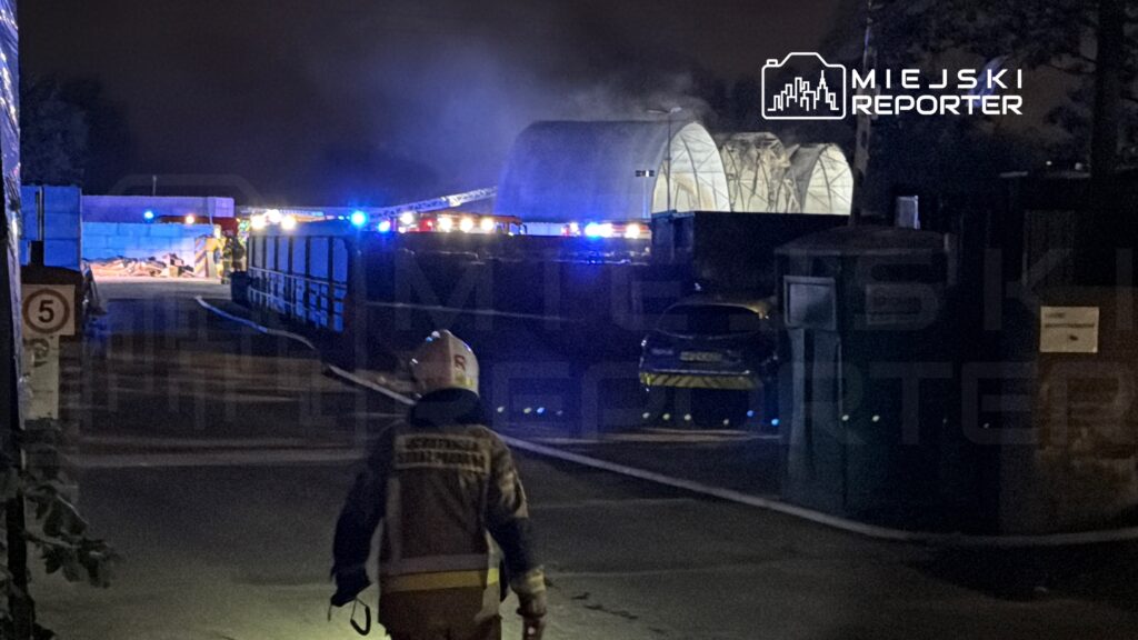 Strażak w hełmie idzie w kierunku miejsca zdarzenia, gdzie widać niebieskie światła radiowozów i dym.