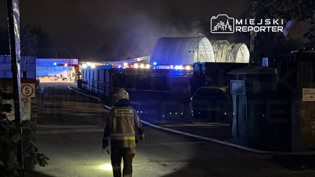 Strażak w hełmie i kamizelce ochronnej przemieszcza się w kierunku miejsca pożaru, otoczonego przez pojazdy ratunkowe.