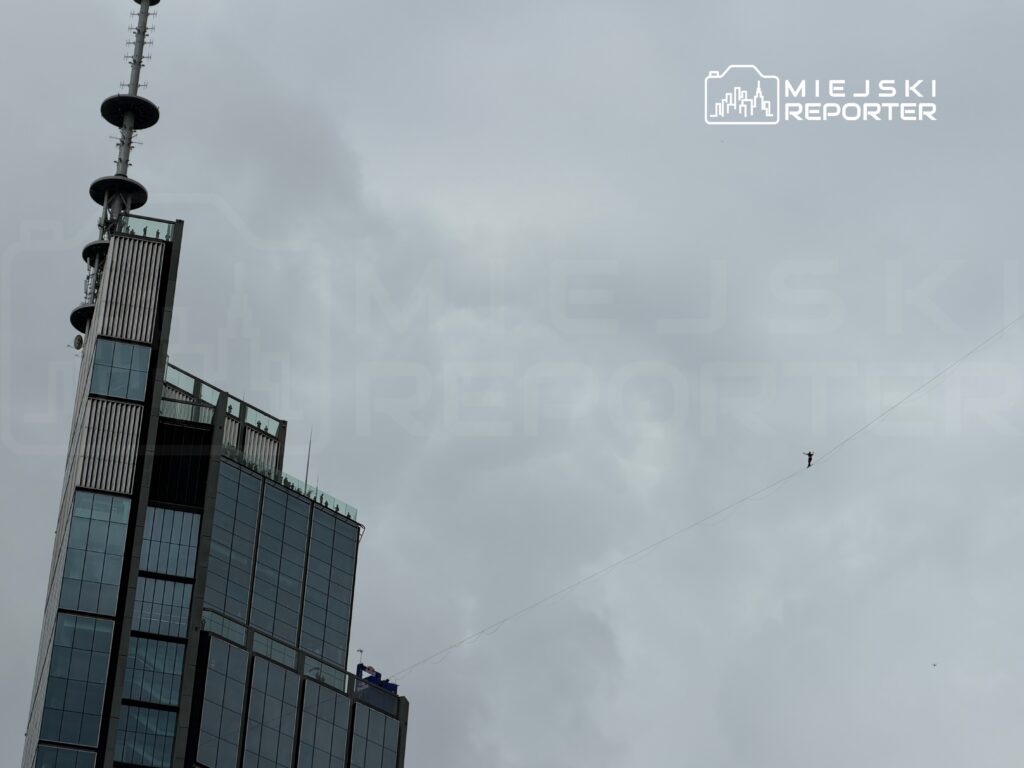 Mężczyzna balansujący na linie rozpiętej między budynkami w szarym, pochmurnym niebie.