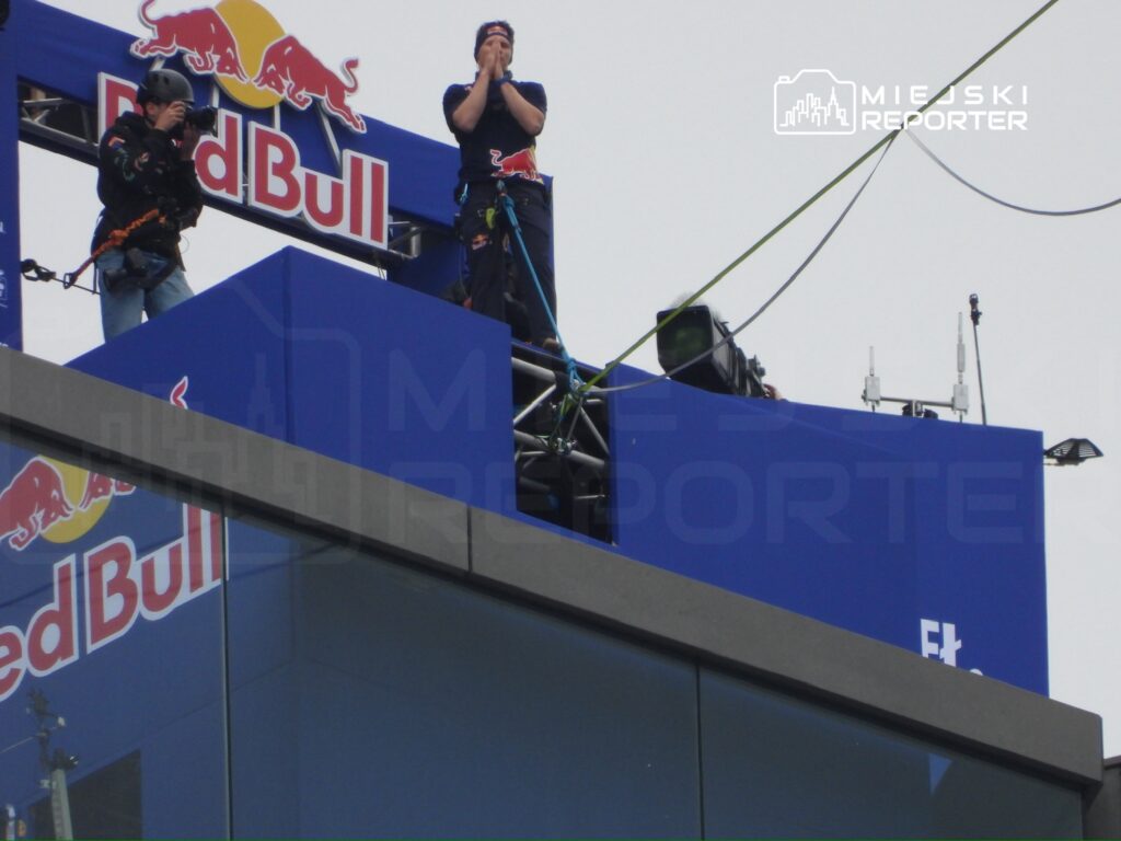 Mężczyzna w kasku stojący na krawędzi dachu, trzymający się za głowę, podczas wydarzenia zorganizowanego przez Red Bull. Obok niego fotograf z aparatem.