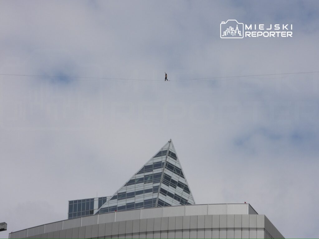 Mężczyzna balansujący na linie między budynkami pod chmurami, z widokiem na nowoczesną architekturę.