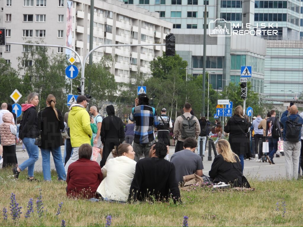 Grupa ludzi stoi i siedzi na trawie, w tle nowoczesne budynki oraz znaki drogowe w miejskiej scenerii.