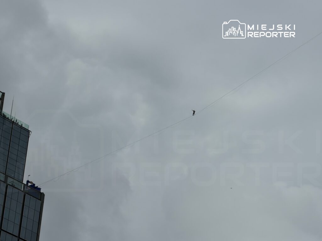 Osoba spacerująca po linie nad miastem, na tle pochmurnego nieba i nowoczesnego budynku.