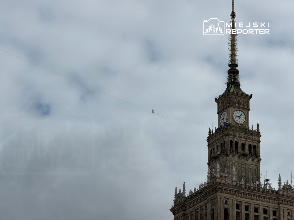 Mężczyzna balansujący na linie między wieżą a budynkiem w chmurzystym niebie.