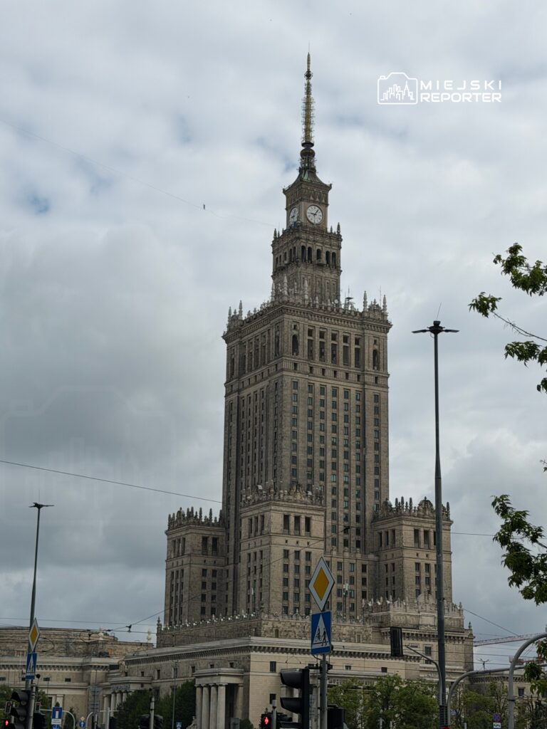 Wysoki, zabytkowy budynek z wieżą zegarową i anteną, otoczony ulicami i sygnalizacją świetlną.