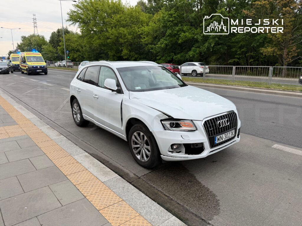 Biały samochód osobowy marki Audi z uszkodzoną przednią częścią stoi na jezdni, obok przejeżdżają karetki pogotowia.