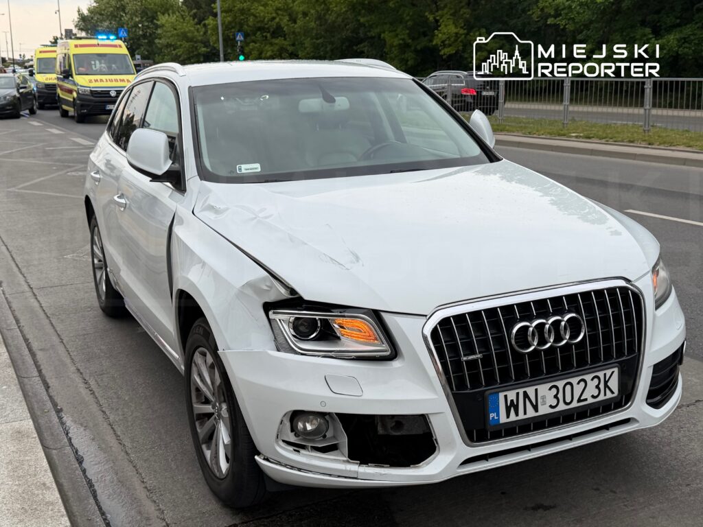 Biały samochód osobowy marki Audi z uszkodzeniami na przednim prawym boku stoi na jezdni, obok widać karetkę pogotowia.