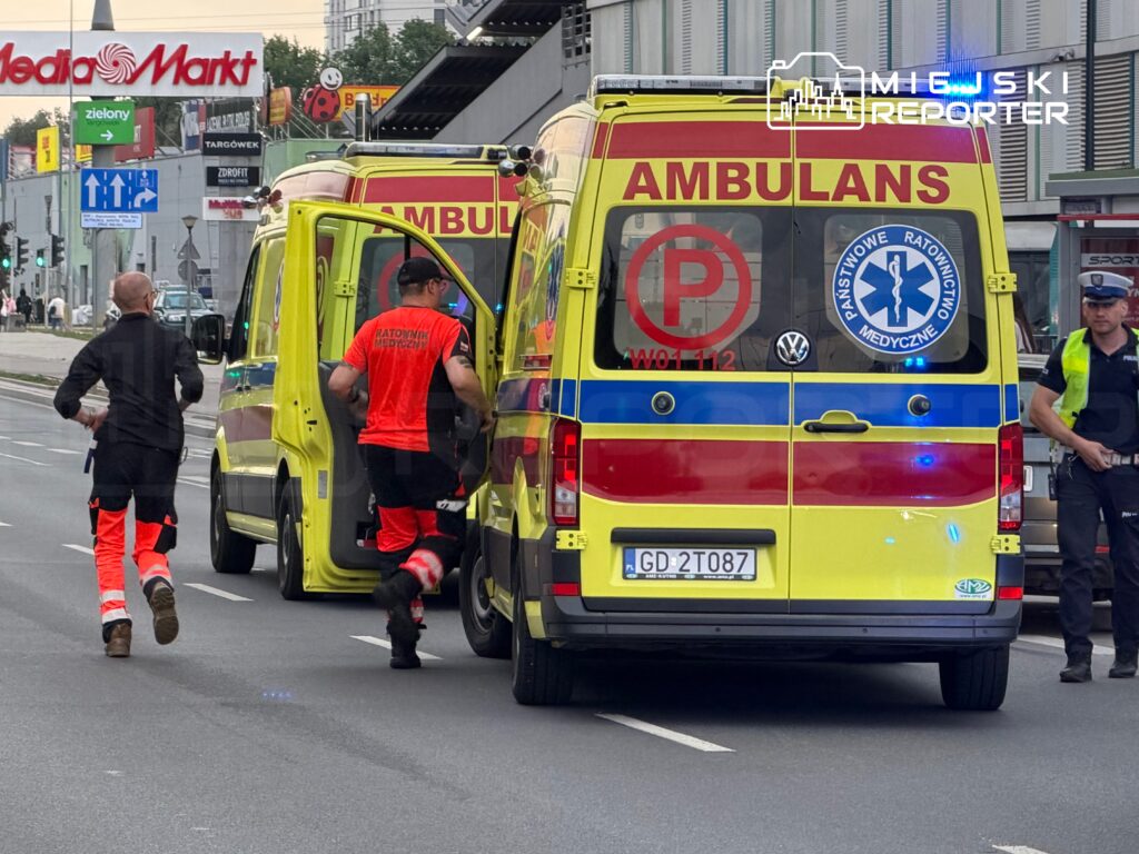 Dwaj ratownicy medyczni w odzieży ochronnej podbiegają do karetki na ulicy, obok widać policyjnego funkcjonariusza.
