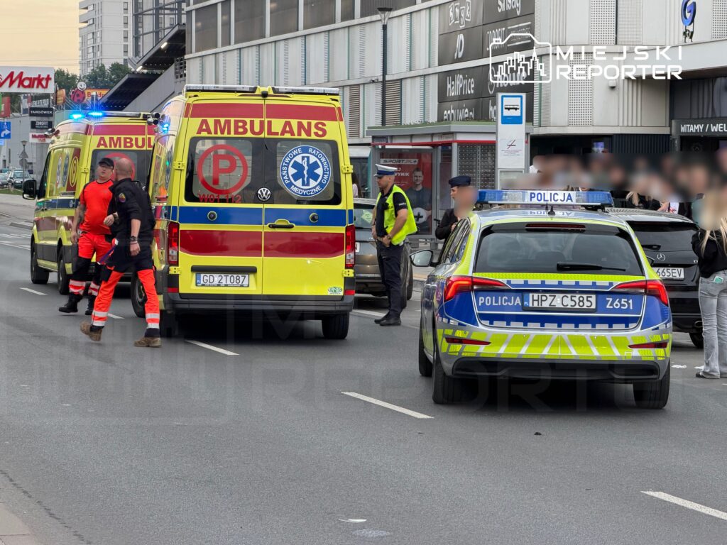 Na ulicy zatrzymane są dwa pojazdy: karetka pogotowia z włączonymi sygnałami oraz radiowóz policji. Ratownicy medyczni rozmawiają z policjantami. W tle widoczny jest budynek handlowy.