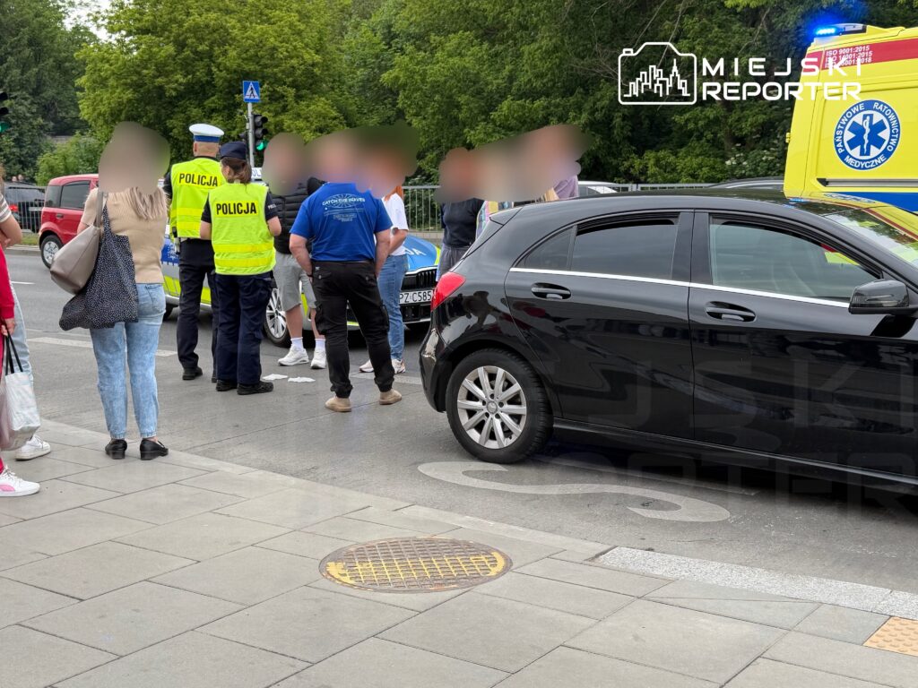 Grupa osób rozmawia z policjantami na ulicy obok zaparkowanego radiowozu i karetki pogotowia.