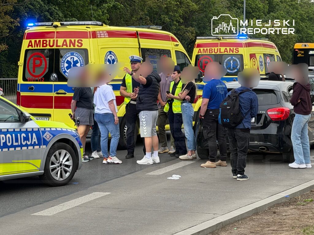 Grupa osób rozmawia z policjantem obok karetki pogotowia i radiowozu na ruchliwej ulicy.