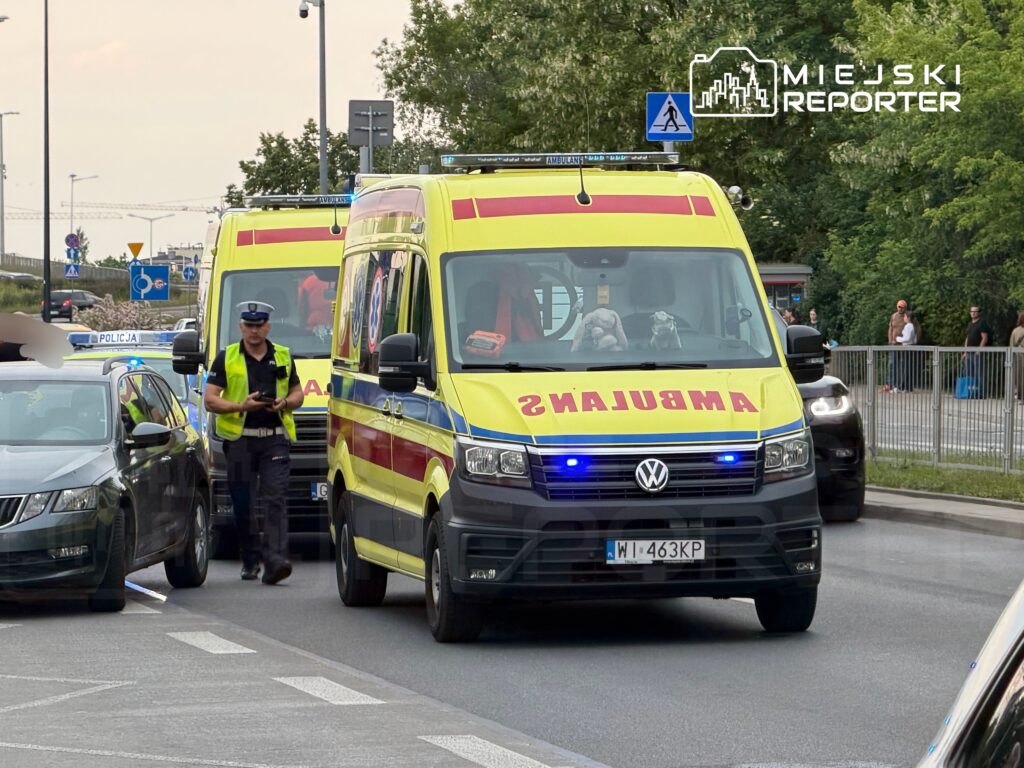 Ambulans z niebieskimi światłami zatrzymany na drodze, obok policjant w kamizelce odblaskowej, w tle inne pojazdy.