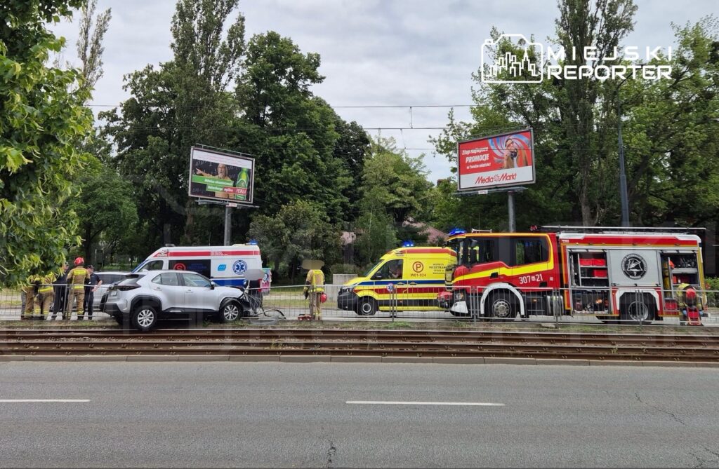 Zespół ratunkowy i straż pożarna pracują przy wypadku drogowym obok torów tramwajowych, w tle billboardy.