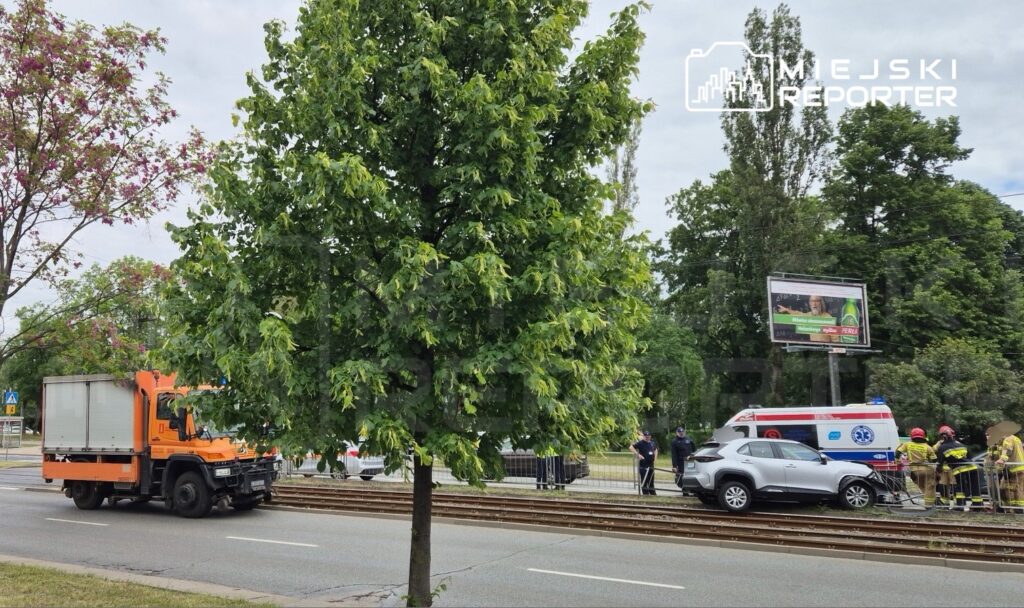 W pobliżu torów tramwajowych stoi uszkodzony samochód, obok niego pracują strażacy i ratownicy medyczni.
