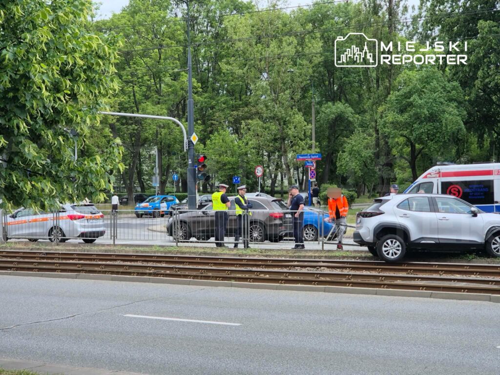 Grupa osób w odblaskowych kamizelkach rozmawia obok zablokowanej drogi, na której stoją samochody i karetka.