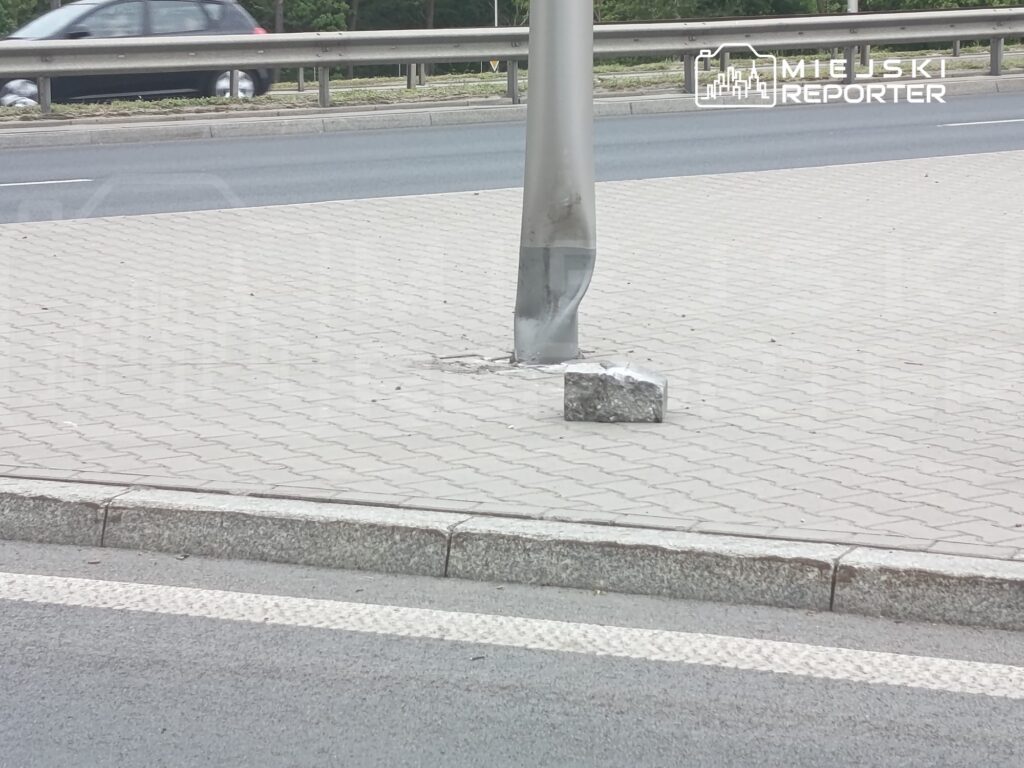 Wielki słup uliczny z uszkodzoną podstawą stoi na chodniku obok ruchliwej drogi, obok leży kamień.