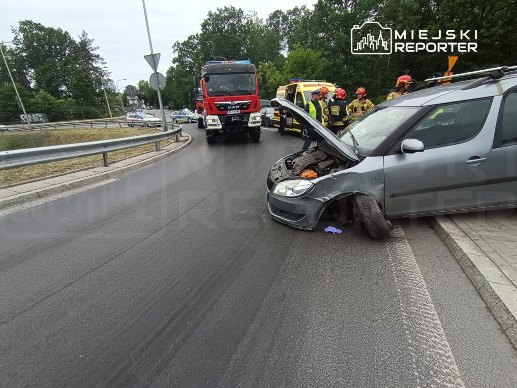 Uszkodzony samochód osobowy na poboczu drogi, obok interweniujący strażacy i karetka.