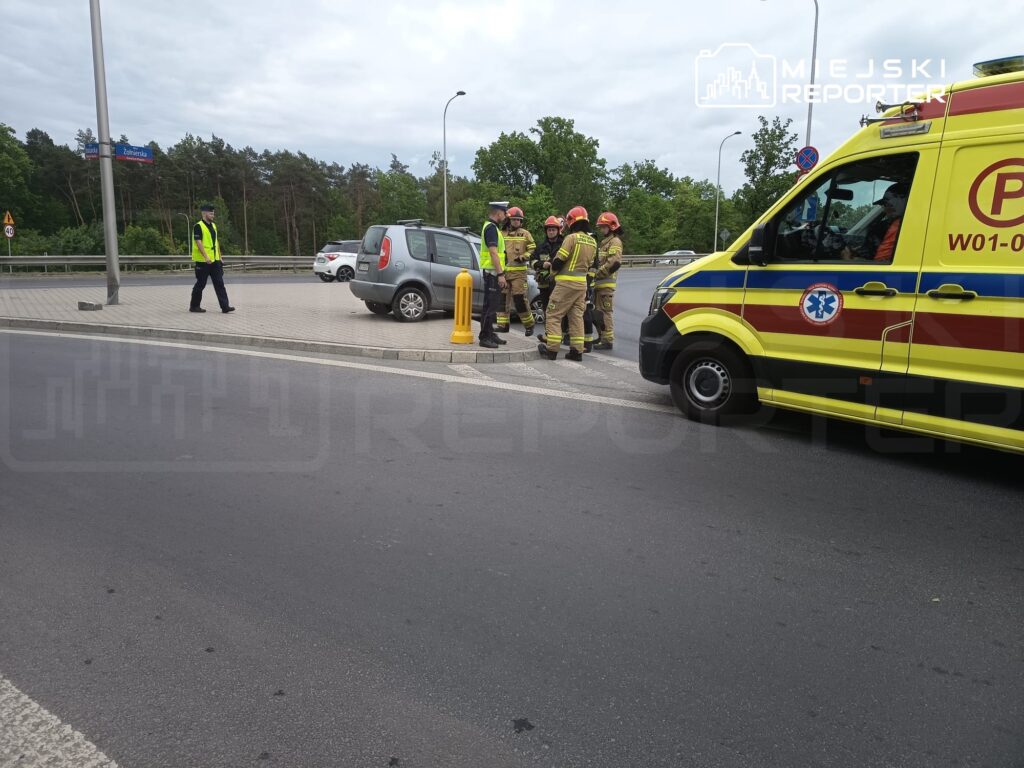 Grupa strażaków rozmawia z ratownikami medycznymi obok karetki na poboczu drogi, w tle widać samochód osobowy.