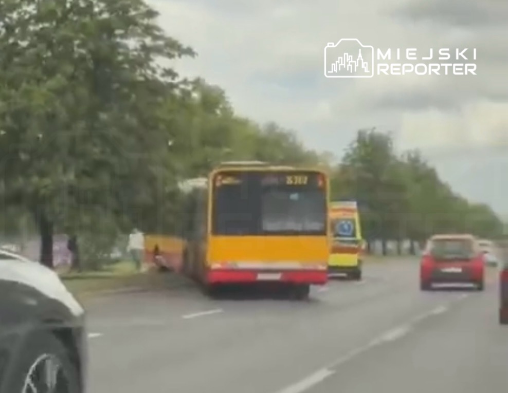 Żółty autobus komunikacji miejskiej zatrzymany na drodze, obok przejeżdżające samochody, w tle drzewa.