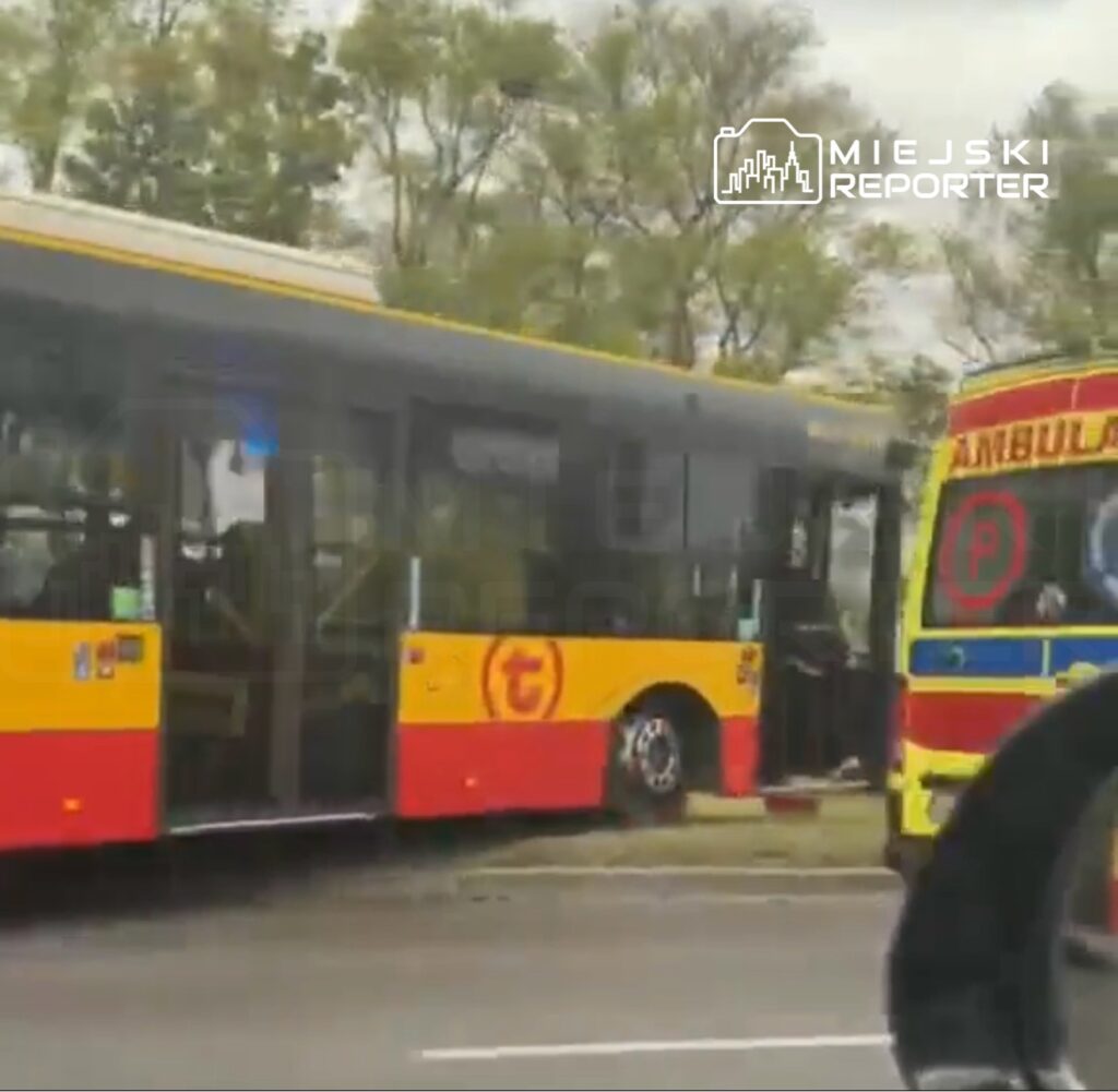 Autobus miejski z otwartymi drzwiami zatrzymany przy jezdni obok karetki pogotowia. W tle drzewa.