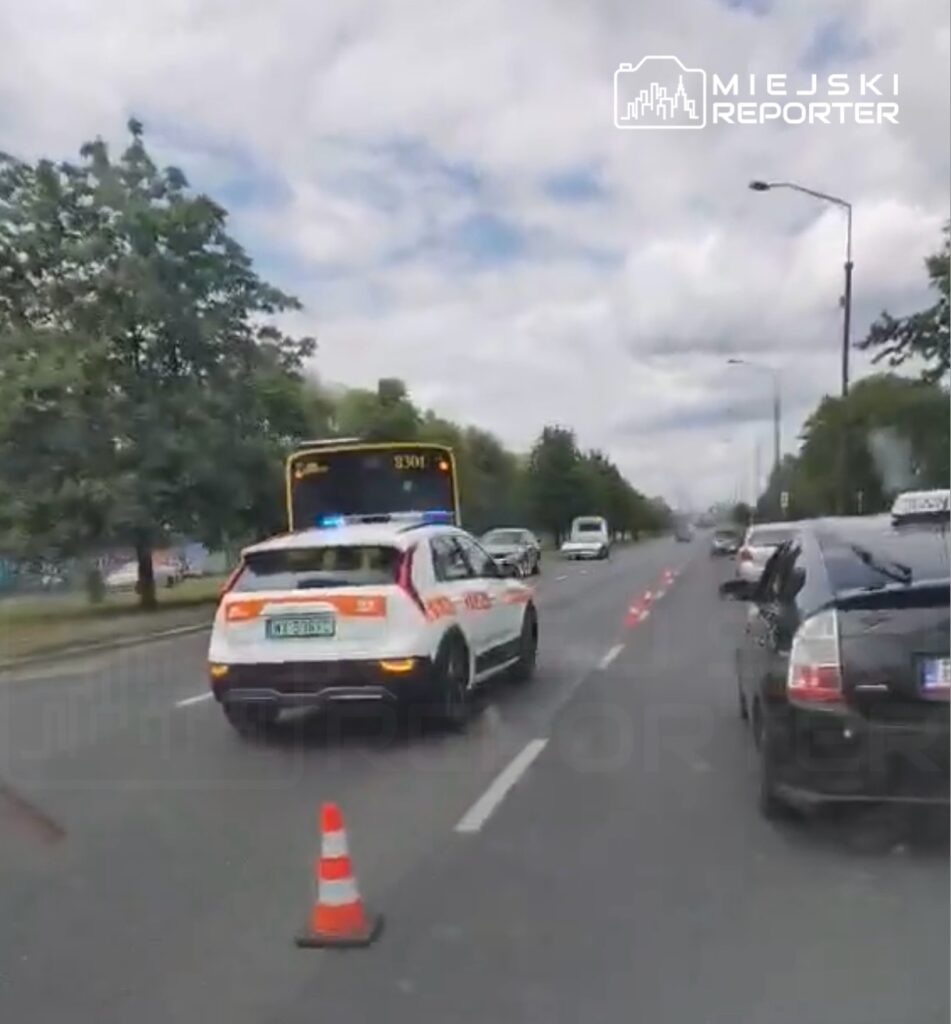 Radiowóz policyjny z włączonymi sygnałami świetlnymi zatrzymuje ruch na drodze, obok widoczny autobus.