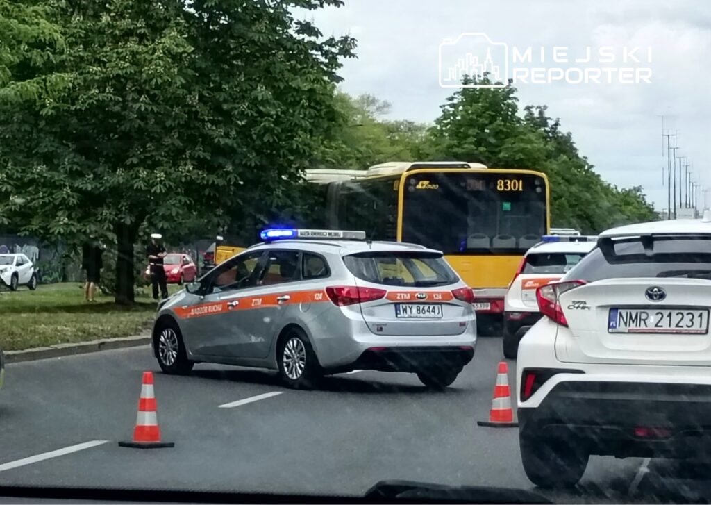 Radiowóz z włączonym sygnałem świetlnym blokuje ruch na drodze, a kierowcy oczekują w zatorze. W tle widoczny autobus.