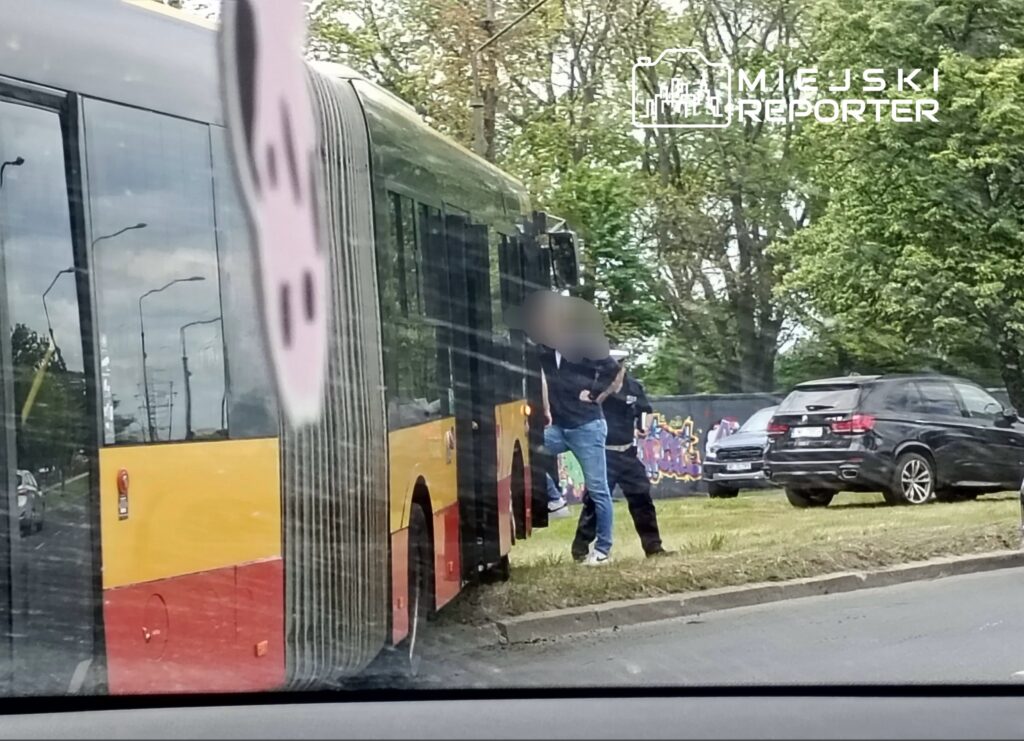 Mężczyzna w dżinsach i ciemnej bluzie zatrzymany przez policjanta obok przystanku autobusowego.