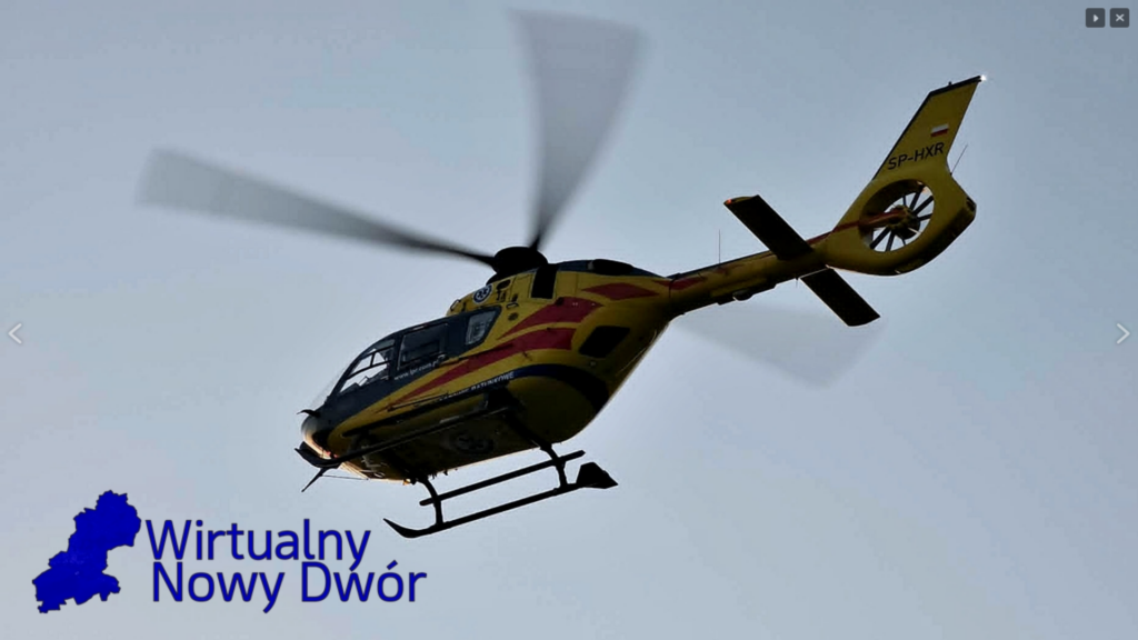 Żółty helikopter z wirującymi wirnikami przelatuje na tle jasnego nieba.